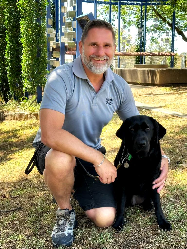 Smiling Aaron kneels next to a black labrador